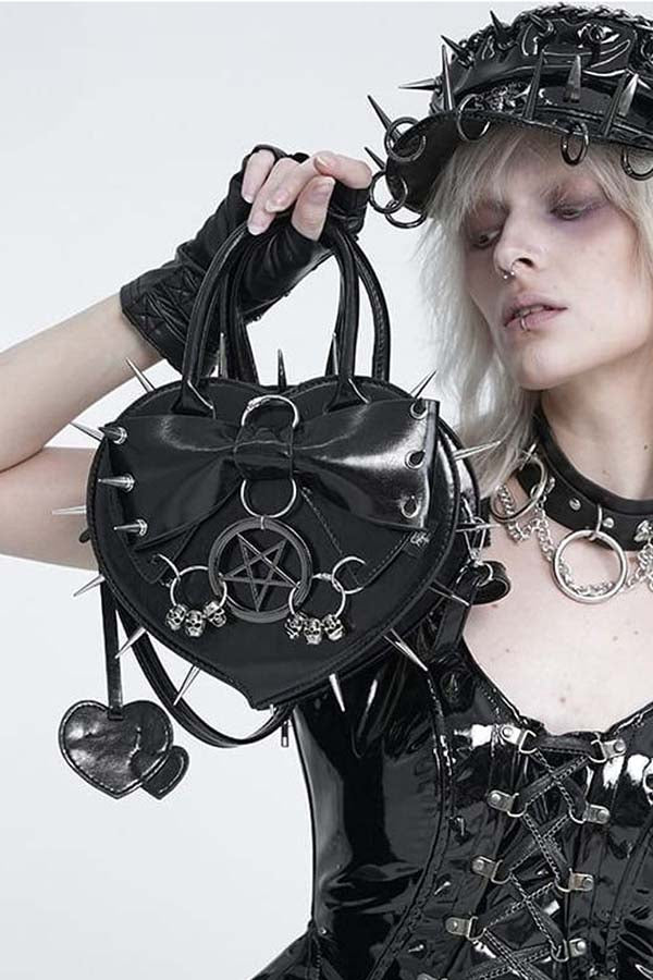 A gothic-style model wearing a black spiked headpiece, black gloves, and a black lace-up top holds up a black heart-shaped leather handbag decorated with metal rings, spikes, pentagrams, and dangling charms against a plain white background.